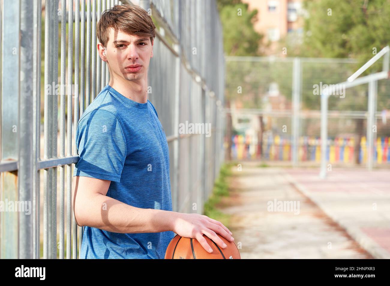 giovane sportivo con una pallacanestro in mano appoggiata contro una recinzione metallica su un campo di pallacanestro Foto Stock