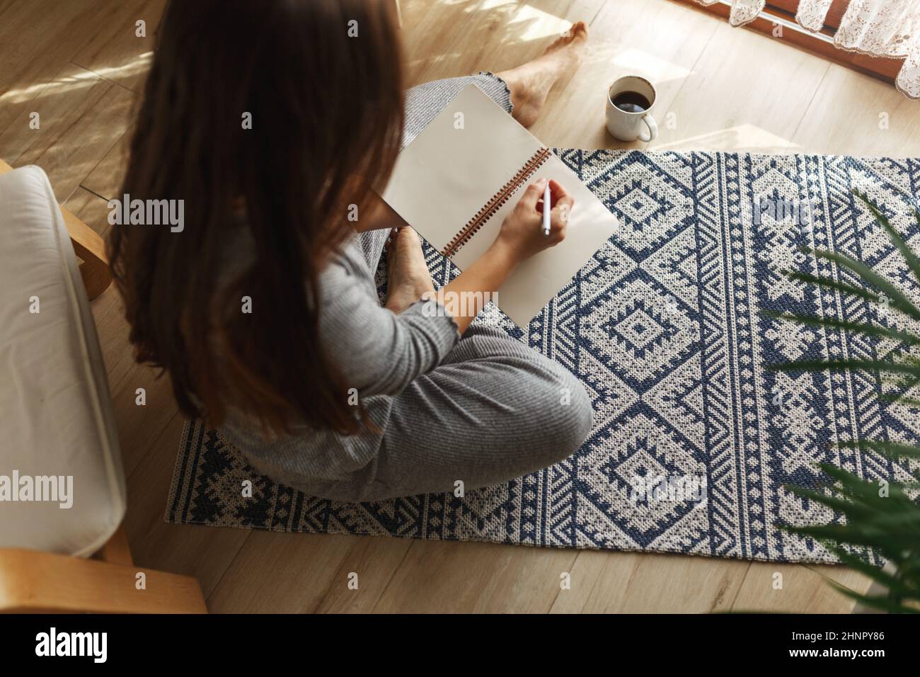 Giovane donna che scrive le pagine del mattino in diario, progettando la sua attività al mattino con il notebook e la penna nelle sue mani. Bere caffè interni casa moderna e luminosa sullo sfondo. Foto Stock