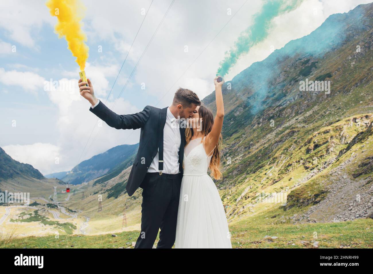 Ritratto di un sposo novellino con il fumo giallo e blu nelle mani che si posa mentre bacia nella montagna. Paesaggio naturale. Giorno del matrimonio. Foto Stock