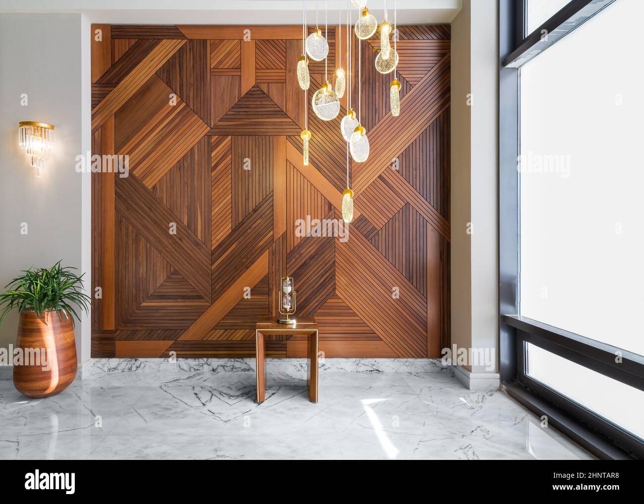 Parete rivestita in legno, alto piantatore con cespugli, moderno lampadario alto in vetro e pavimento in marmo bianco Foto Stock