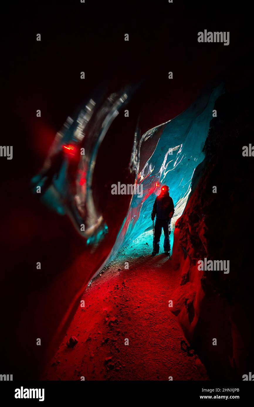 Silhouette di Alpinista in casco con lampada a incandescenza rossa in una profonda e stretta grotta di ghiaccio glaciale con luci ciano esplorare il ghiacciaio invernale di montagna in Kazakhstan Foto Stock