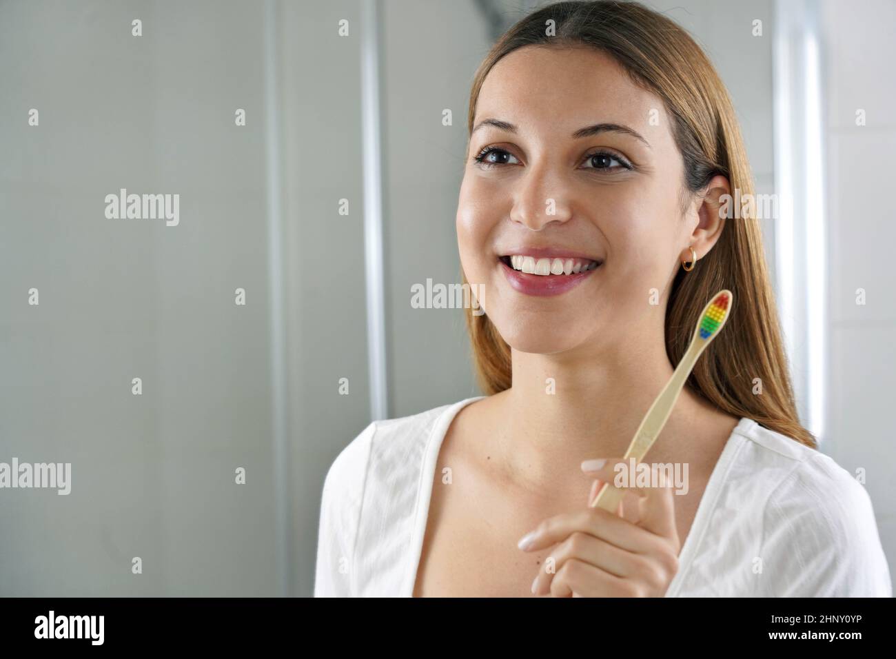 Giovane donna sorridente tiene uno spazzolino di bambù che si guarda nello specchio di casa. Cura dentale, spazzolino ecologico di bambù, nessun spreco di li Foto Stock