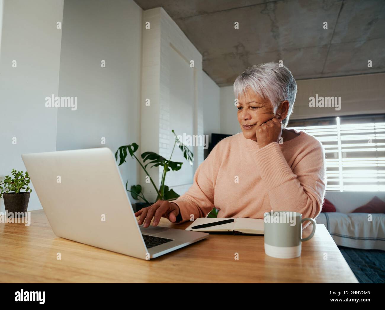 Anziana donna multietnica preoccupata mentre scrollando il Internet sul suo laptop. Sedersi al banco della cucina a casa. Foto Stock