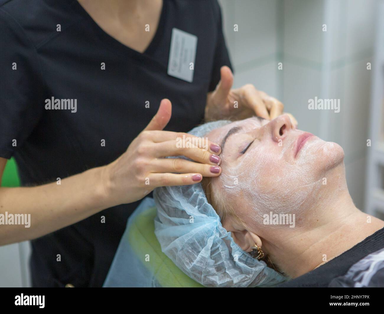 Una donna adulta di 63 anni si trova sulla procedura di ringiovanimento al beautician con una maschera sul suo viso. Le mani del cosmetologo massaggiano l'aragosta. Ciao Foto Stock