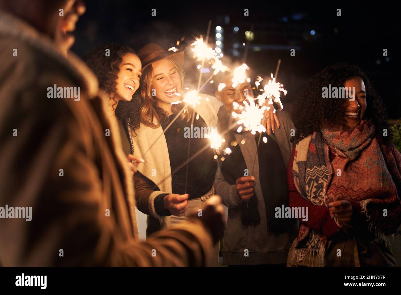 Gruppo multiculturale di amici che ridono mentre illuminano gli scintillanti in una festa alla moda sul tetto Foto Stock