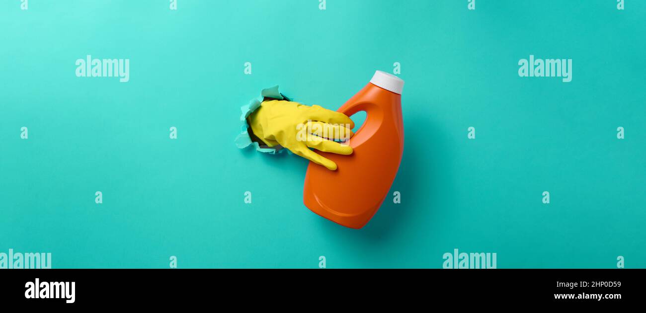 una mano in un guanto di pulizia in lattice giallo tiene una bottiglia arancione di plastica di detergente liquido contro uno sfondo blu chiaro. Banner Foto Stock