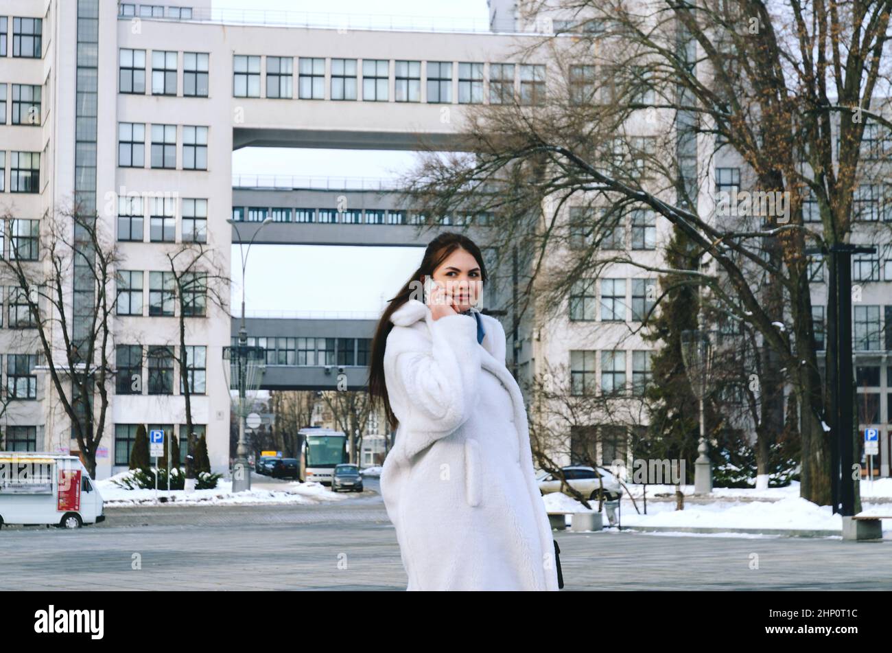 Ritratto urbano di giovane donna elegante in inverno abiti casual, giacca bianca. Camminando nella strada della città, parlando al telefono, facendo selfie Foto Stock