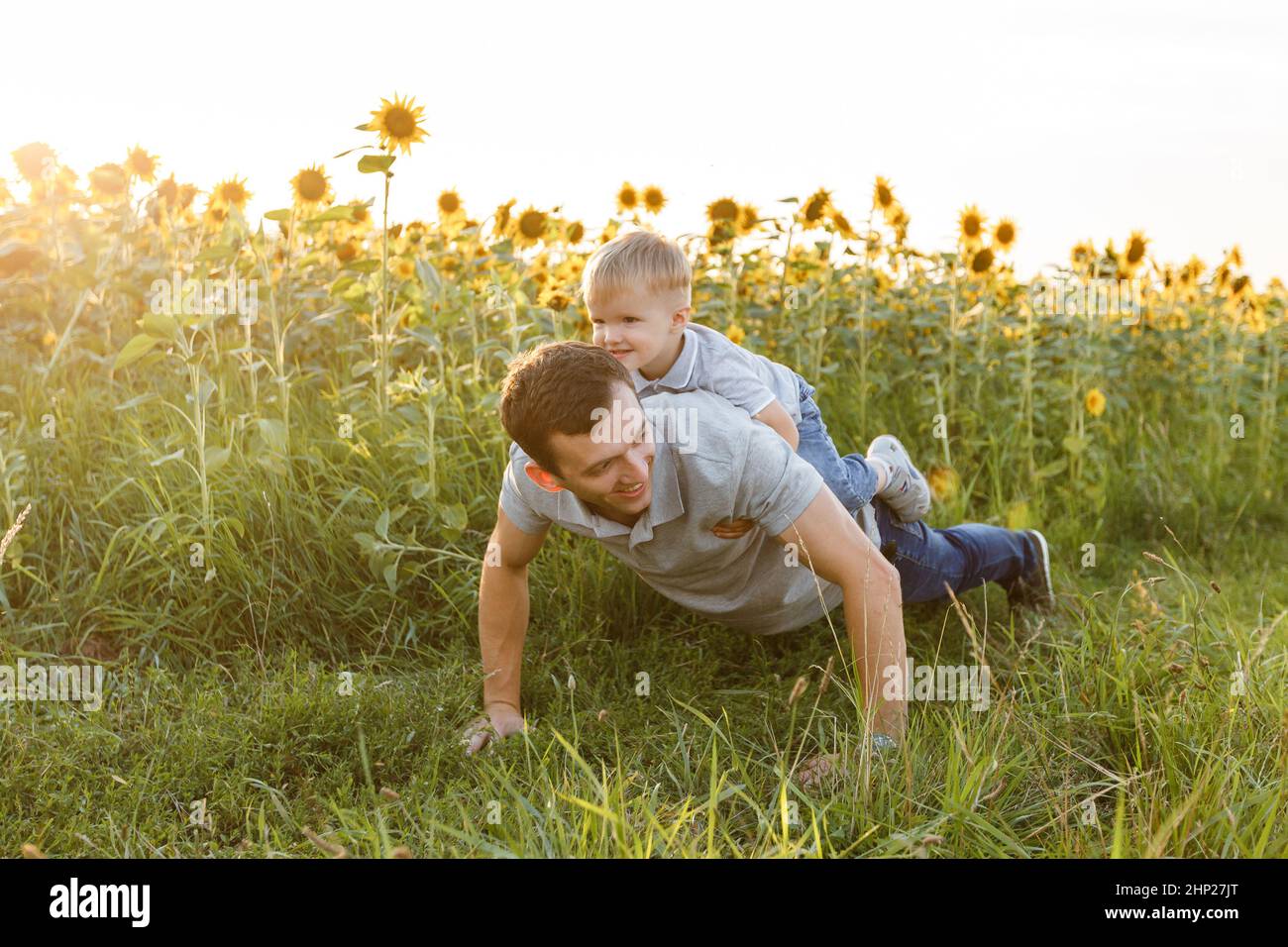 Padre e figlio che fanno spingere insieme in campo di girasoli. Ragazzino seduto sulle spalle del papà e divertito. Stile di vita estivo all'aperto, fami Foto Stock