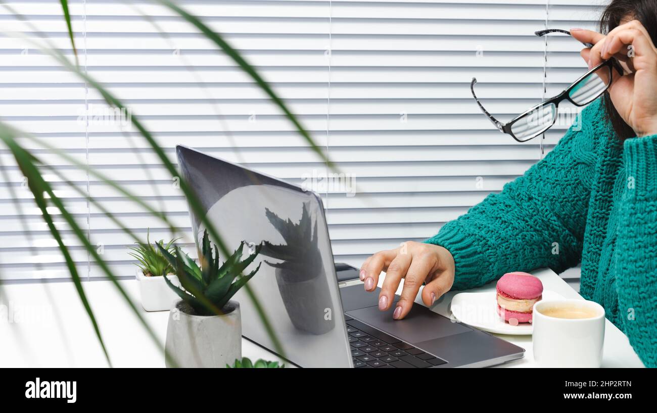 una donna in un maglione blu si siede a un tavolo bianco che lavora a un notebook. Con l'altra mano appoggia la testa, la fatica e il lavoro in eccesso. Burnout nel plac di lavoro Foto Stock