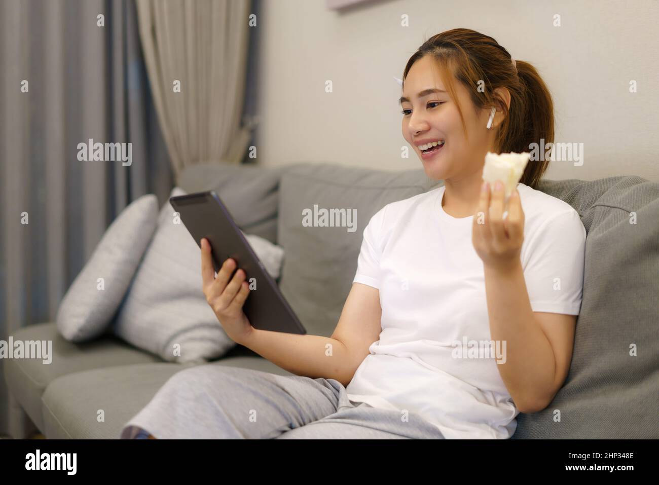 Donna asiatica che lavora e incontra la sua collega di lavoro online in tablet digitale e mangia sandwich sul divano nel salotto di casa di notte. Foto Stock