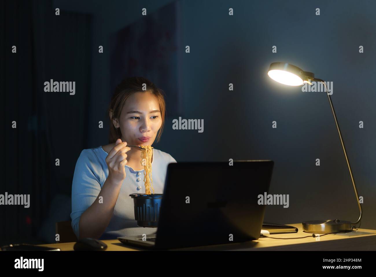 Donna d'affari intelligente freelance asiatica che mangia spaghetti istantanei mentre lavora su un computer portatile nel soggiorno a casa di notte ragazza asiatica felice seduta sopra Foto Stock