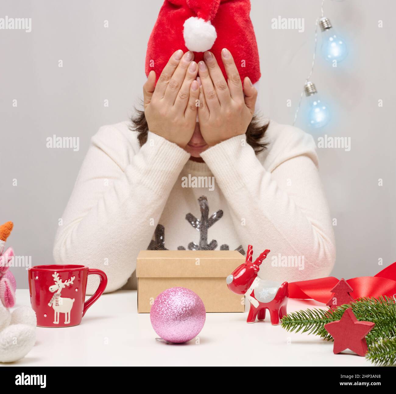 Una donna in un maglione bianco siede al tavolo e avvolge regali per Natale. Tieni la testa con le mani, tristezza Foto Stock