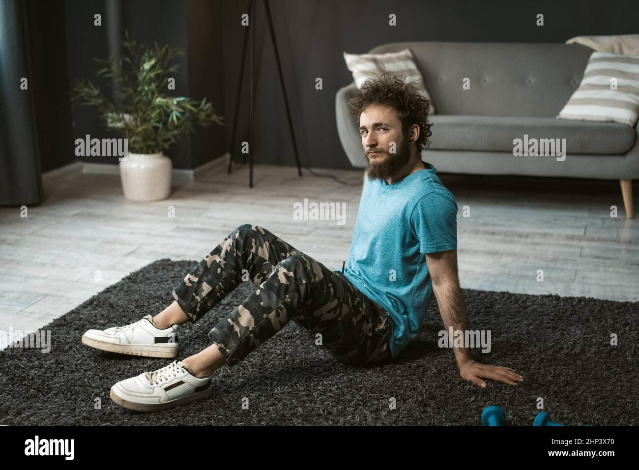Bel giovane uomo che fa a casa allenarsi seduto sul tappeto rilassato a casa guardando la macchina fotografica. Divertente uomo riccio capelli con una barba disordine facendo esercizio a casa. Concetto di vita sana. Foto Stock