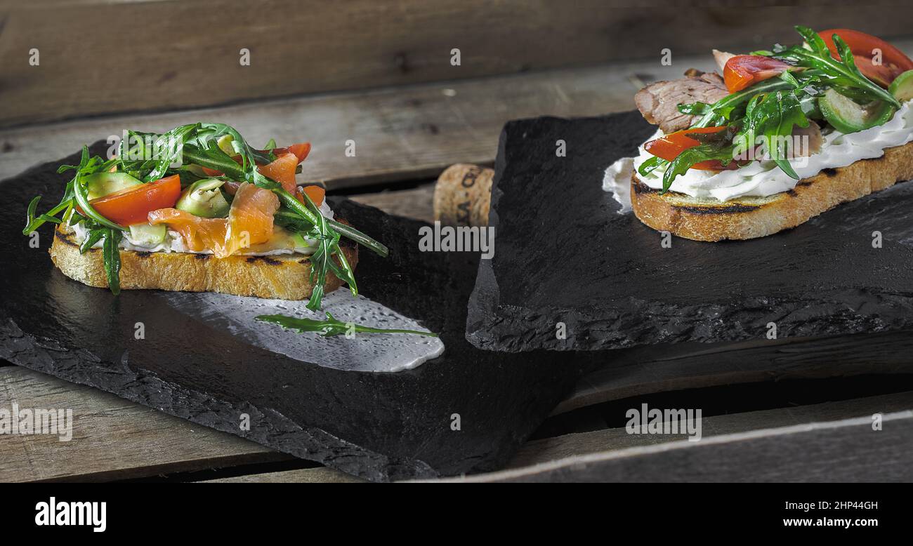 due bruschette con salmone e roast beef, con salsa di panna, pomodori e rucola, giacciono su supporti in pietra su una superficie di legno Foto Stock