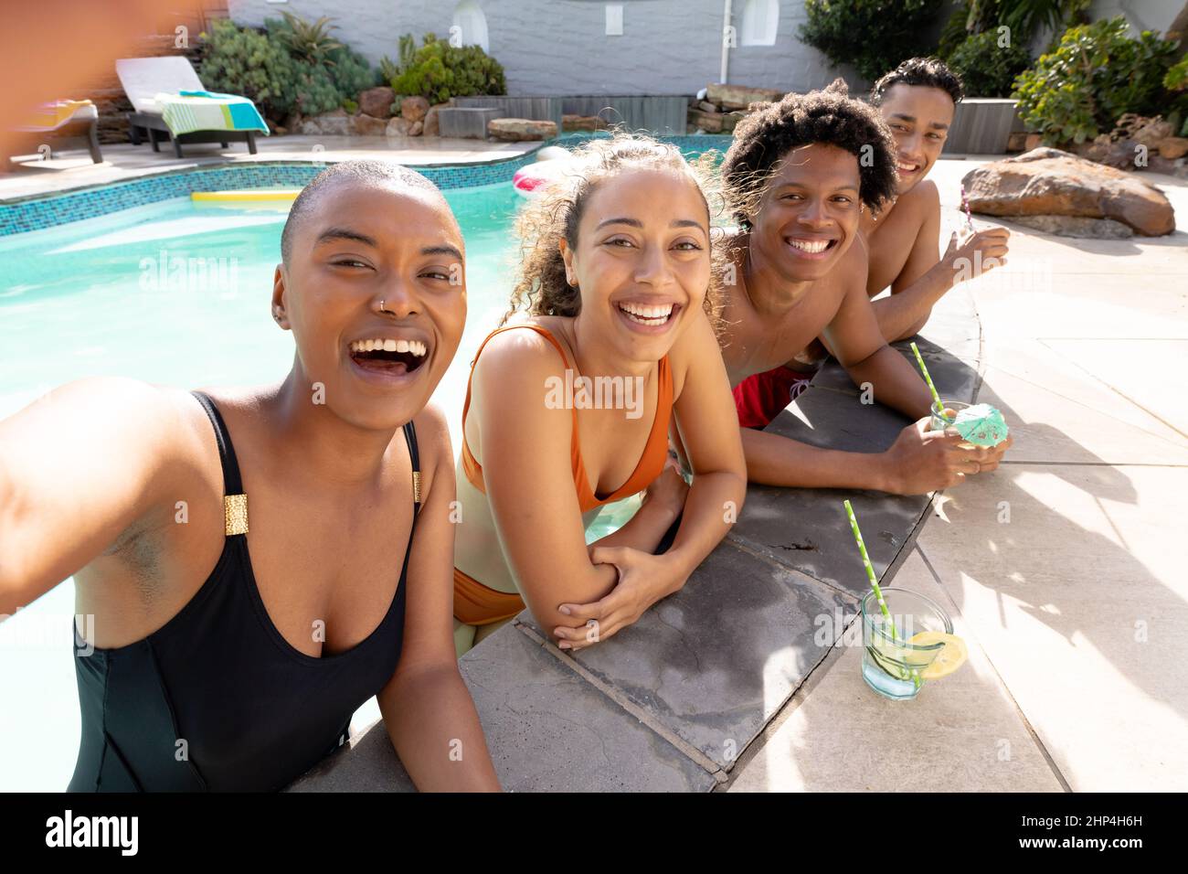Allegra donna multirazziale che prende selfie con gli amici che si appoggiano a bordo piscina durante la giornata di sole Foto Stock