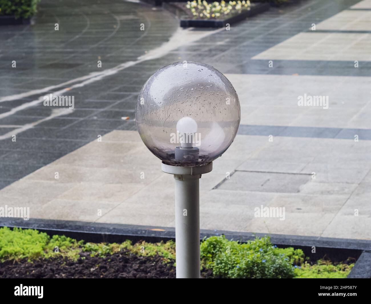 Una lampada da strada rotonda bassa bagnata con una copertura in vetro trasparente in un letto di fiori sullo sfondo di una piazza con piastrelle di marmo Foto Stock