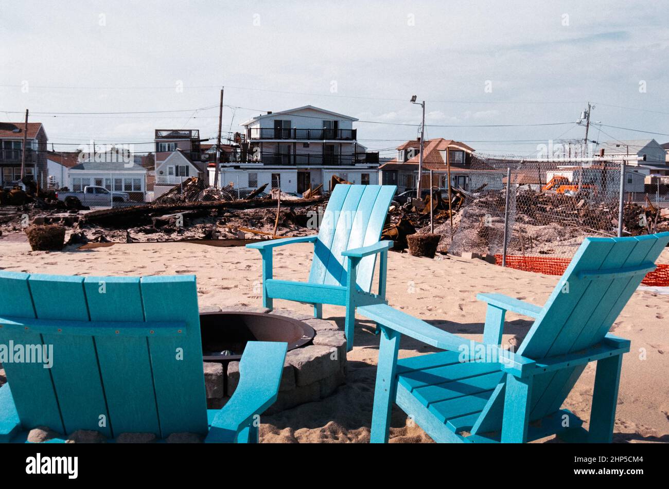 Quattro sedie all'aperto circondano un fuoco di fronte alle rovine di un incendio di allarme del 9 a Salisbury Beach, NH durante l'inverno. Distruzione e macerie circondano Foto Stock