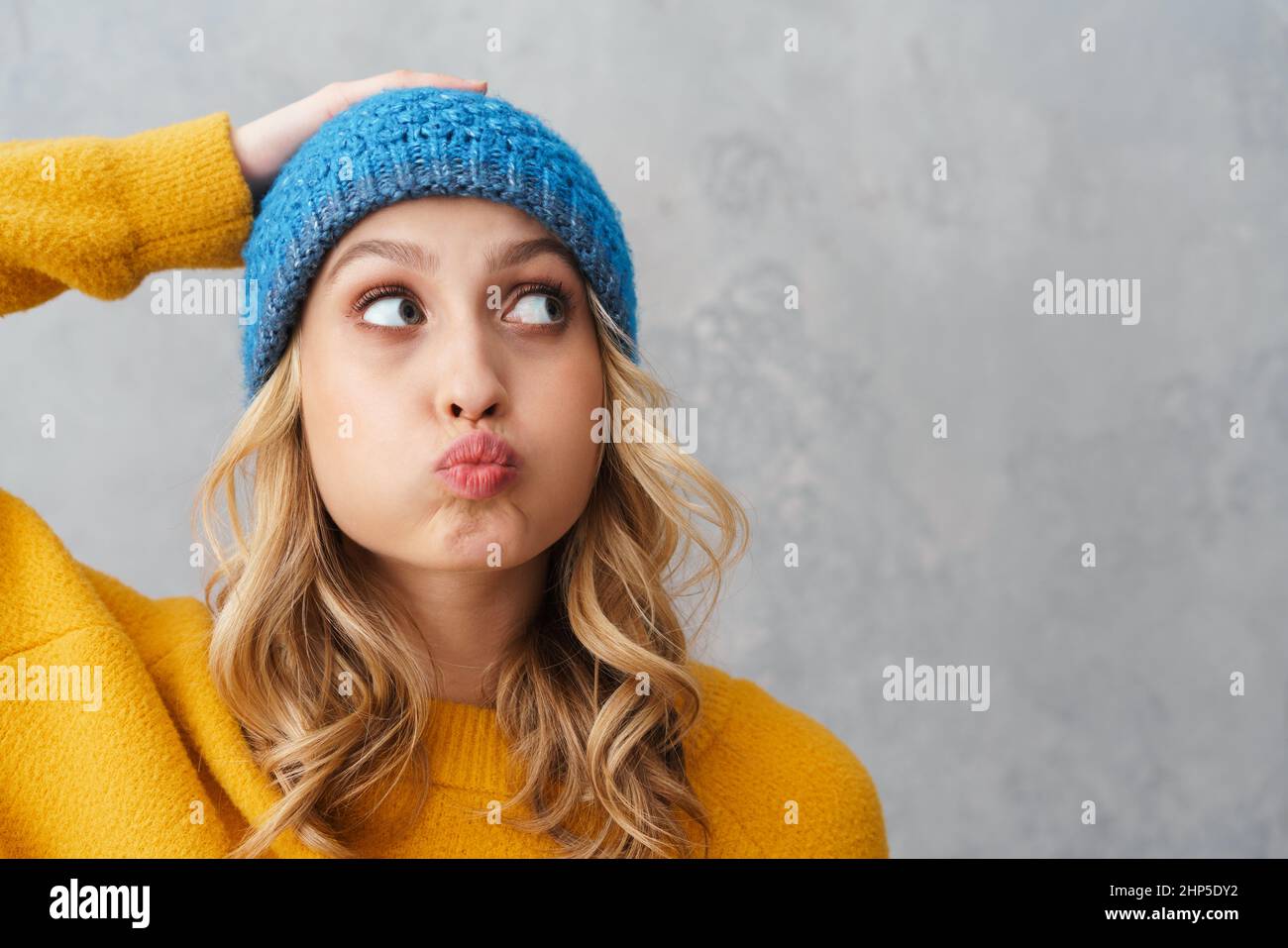 Giovane donna divertente in cappello a maglia che fa baciare le labbra e guardando da parte isolato sopra il muro grigio Foto Stock