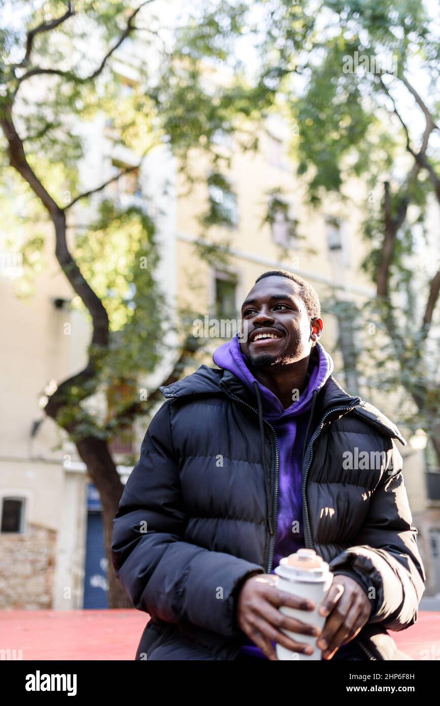 Angolo basso di uomo afro-americano positivo in outerwear guardando via con sorriso. Mentre si gusta il caffè da asporto da zero tazzina rifiuti su sfondo sfocato della strada della città in giorno di sole Foto Stock