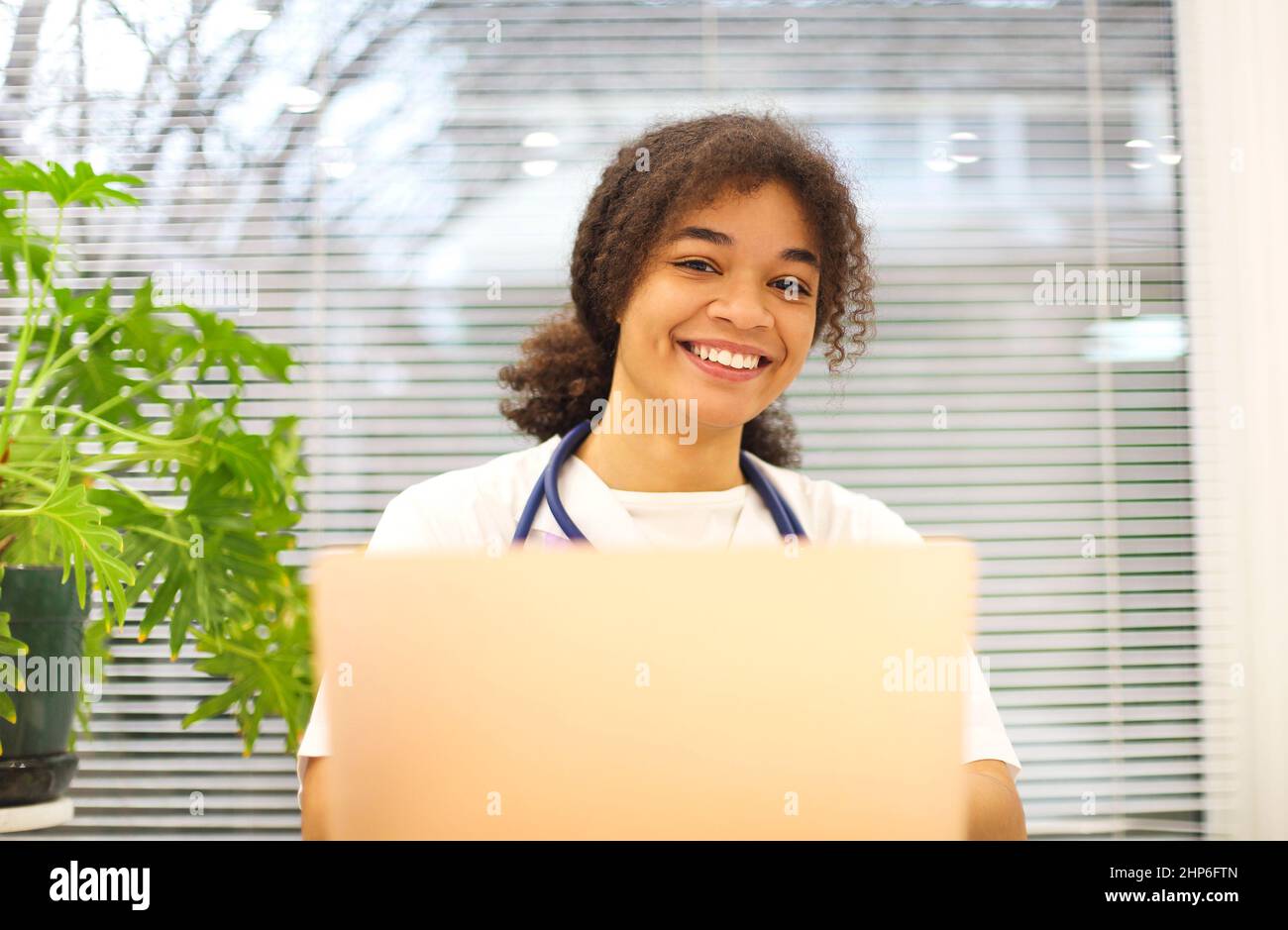 Giovane felice medico afroamericano seduto sul posto di lavoro guardando la macchina fotografica sorridendo mentre si lavora su un computer portatile in clinica moderna, felice etnico Foto Stock