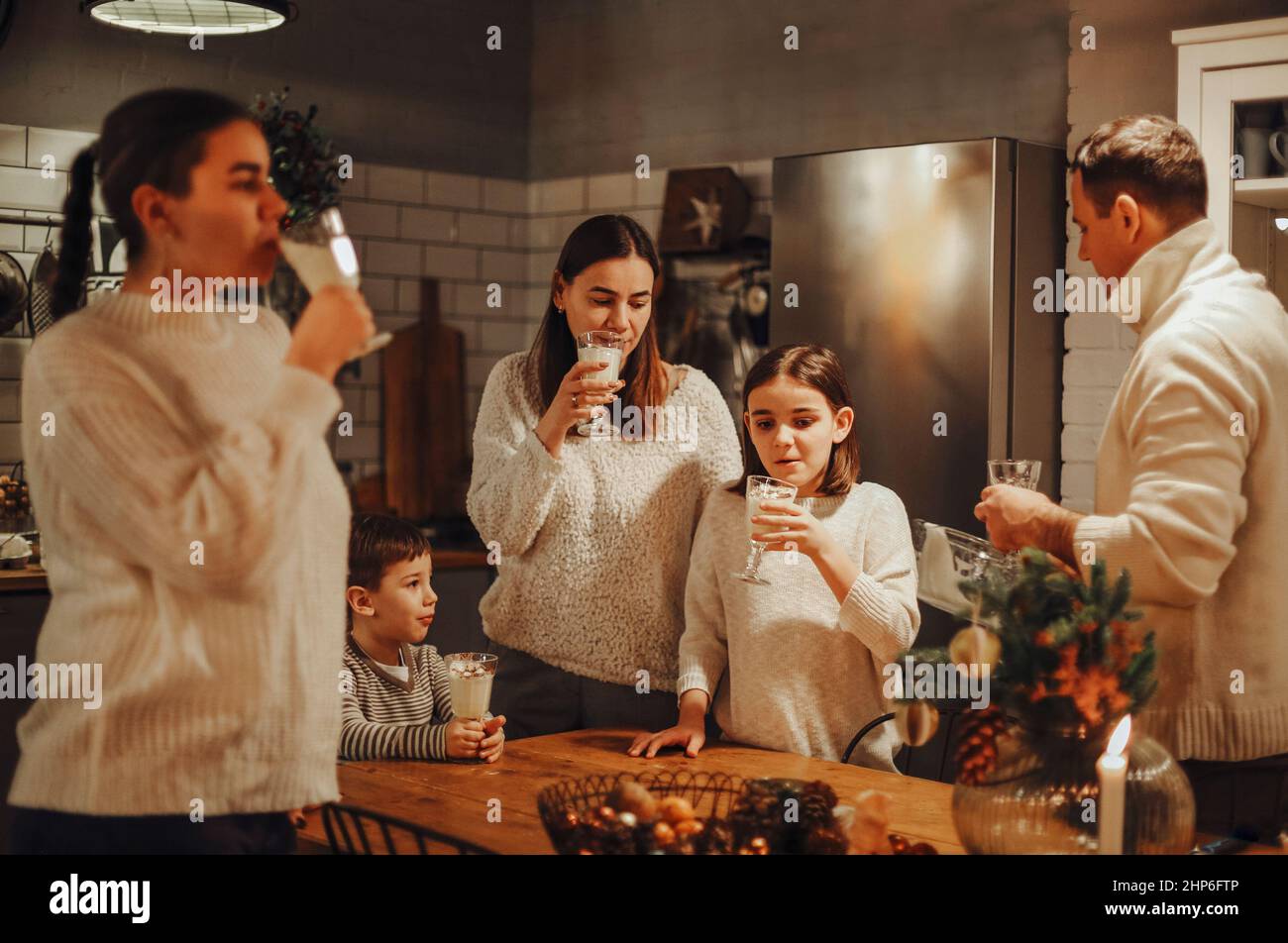 Felice famiglia genitori gioiosi con due bambini che versano egnog fatto in casa in bicchieri, mentre in piedi dietro tavolo di legno insieme e cucinare in accogliente rustico Foto Stock