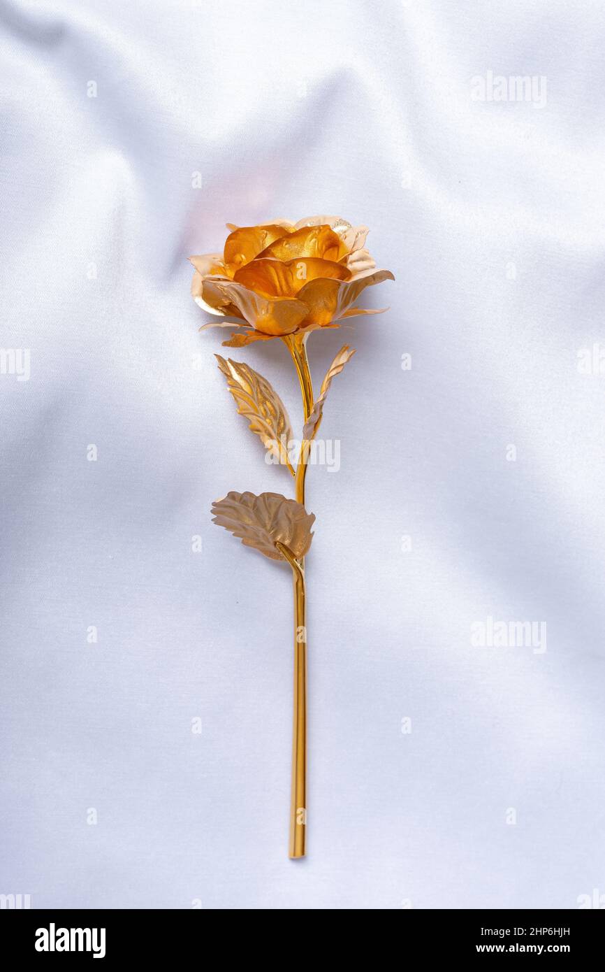 Bel fiore di metallo fatto a mano rosa dorata con foglie e gambo su uno sfondo di seta bianca, vista dall'alto. Gioielleria regalo per una vacanza per una persona cara. STY Foto Stock