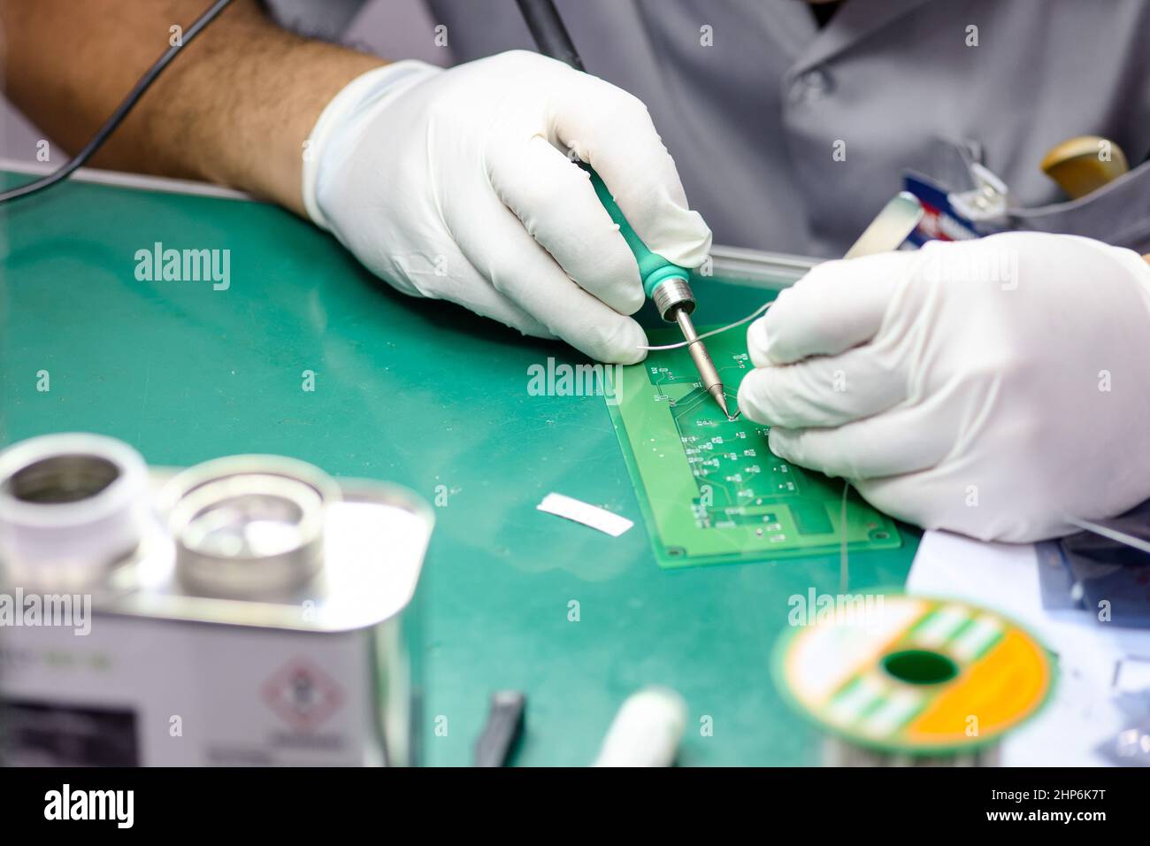 Il maschio del tecnico di primo piano sui guanti di gomma e la tuta da lavoro sta usando la saldatura per saldare la scheda elettronica a circuito stampato sulla scrivania dell'ufficio in fabbrica Foto Stock