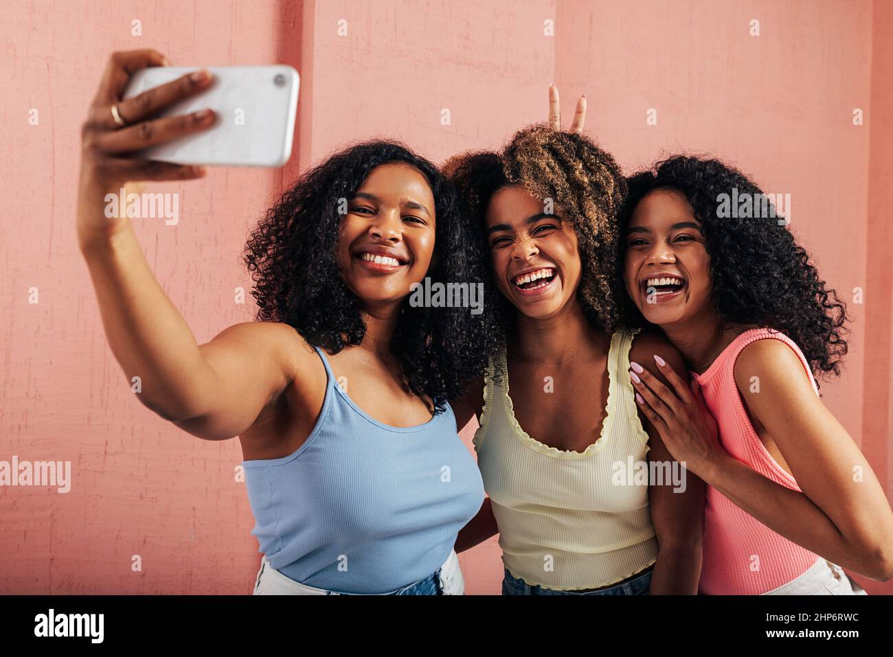 Tre donne con capelli ricci ridendo insieme mentre prendono selfie contro una parete rosa Foto Stock