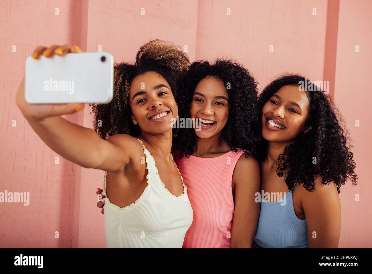 Giovani donne in casuali luminosi in piedi insieme e prendere selfie su un telefono cellulare. Buone femmine che si divertono contro il muro rosa all'aperto. Foto Stock