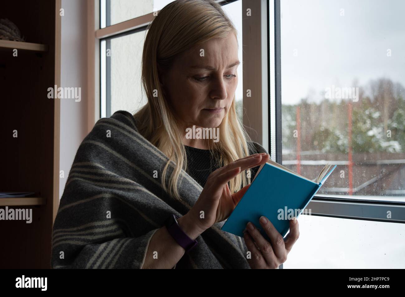 Donna che legge un libro blu brillante alla finestra in inverno vestito di sciarpa di lana Foto Stock
