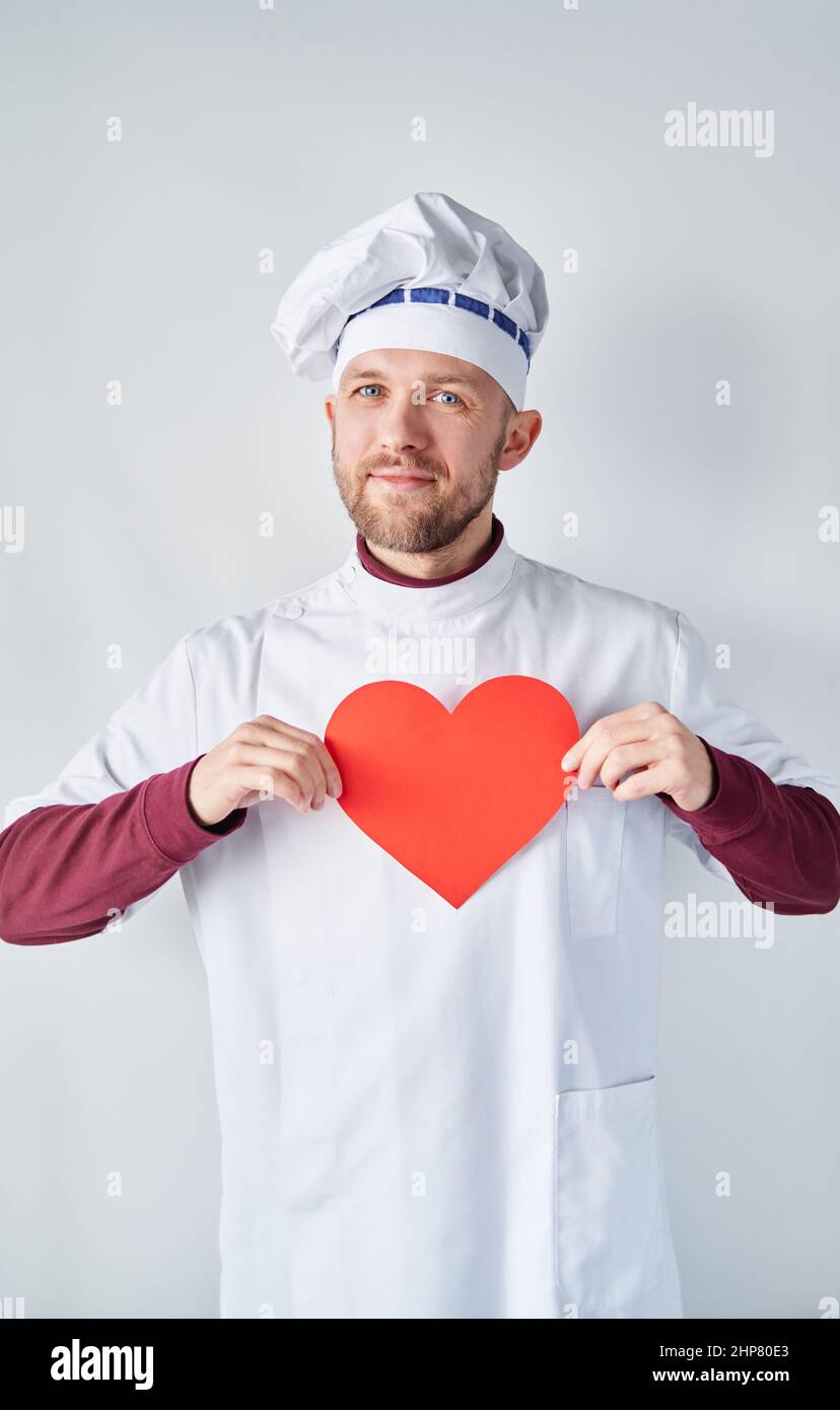 Tema del giorno dello chef: Il fornaio maschile caucasico allegro o lo chef in uniforme bianca con cuore di carta rosso. Ristorante di concetto caffè. Cuoco chef che tiene un cuore rosso di fronte a indumenti da lavoro. Foto di alta qualità Foto Stock