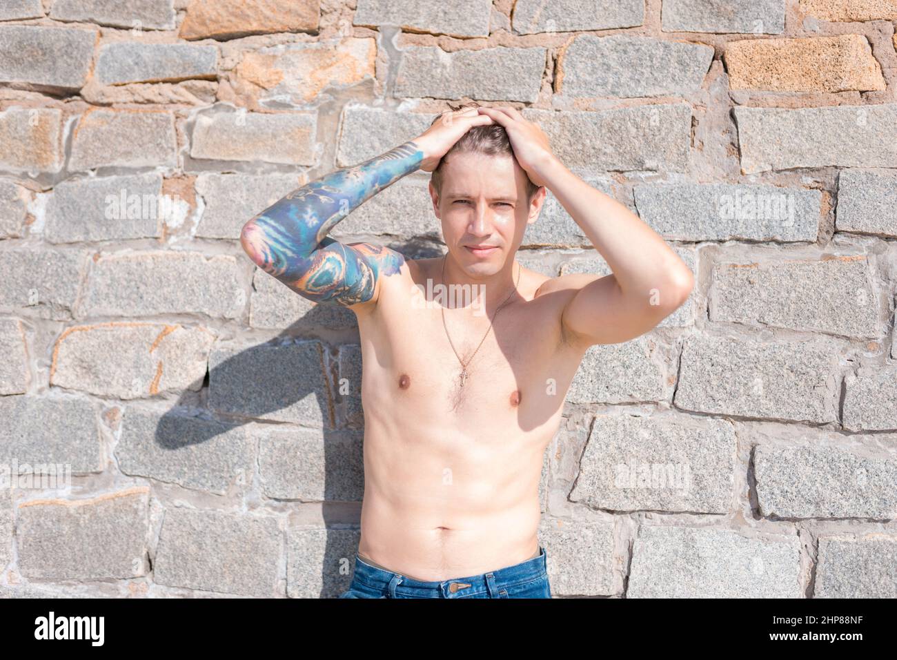 Attraente giovane uomo caucasico nudo-chested con tatuaggio a braccio pieno in piedi contro il muro di pietra. Foto di alta qualità Foto Stock