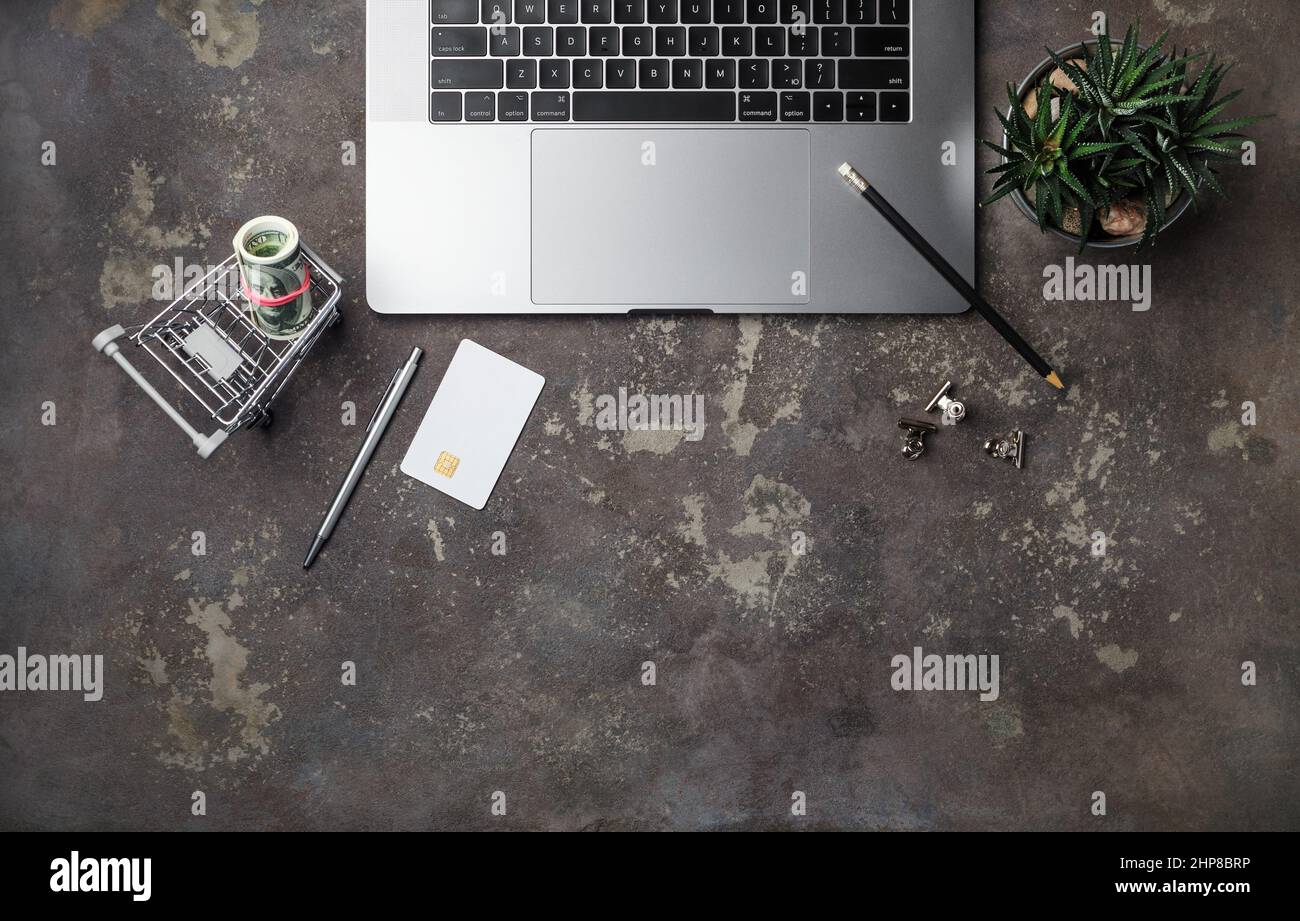 Shopping online e concetto di pagamento. Dollari, carta di credito, carrello, matita, penna succulenta pianta e tastiera laptop su sfondo concreto. Piatto Foto Stock