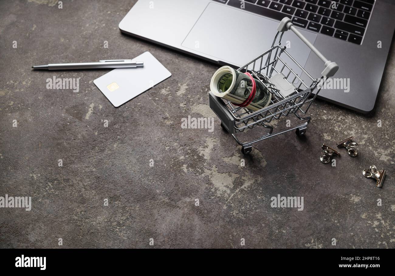 Concetto di shopping in linea del Internet. Computer portatile, carrello con denaro, carta di credito, penna e clip su sfondo di cemento. Foto Stock