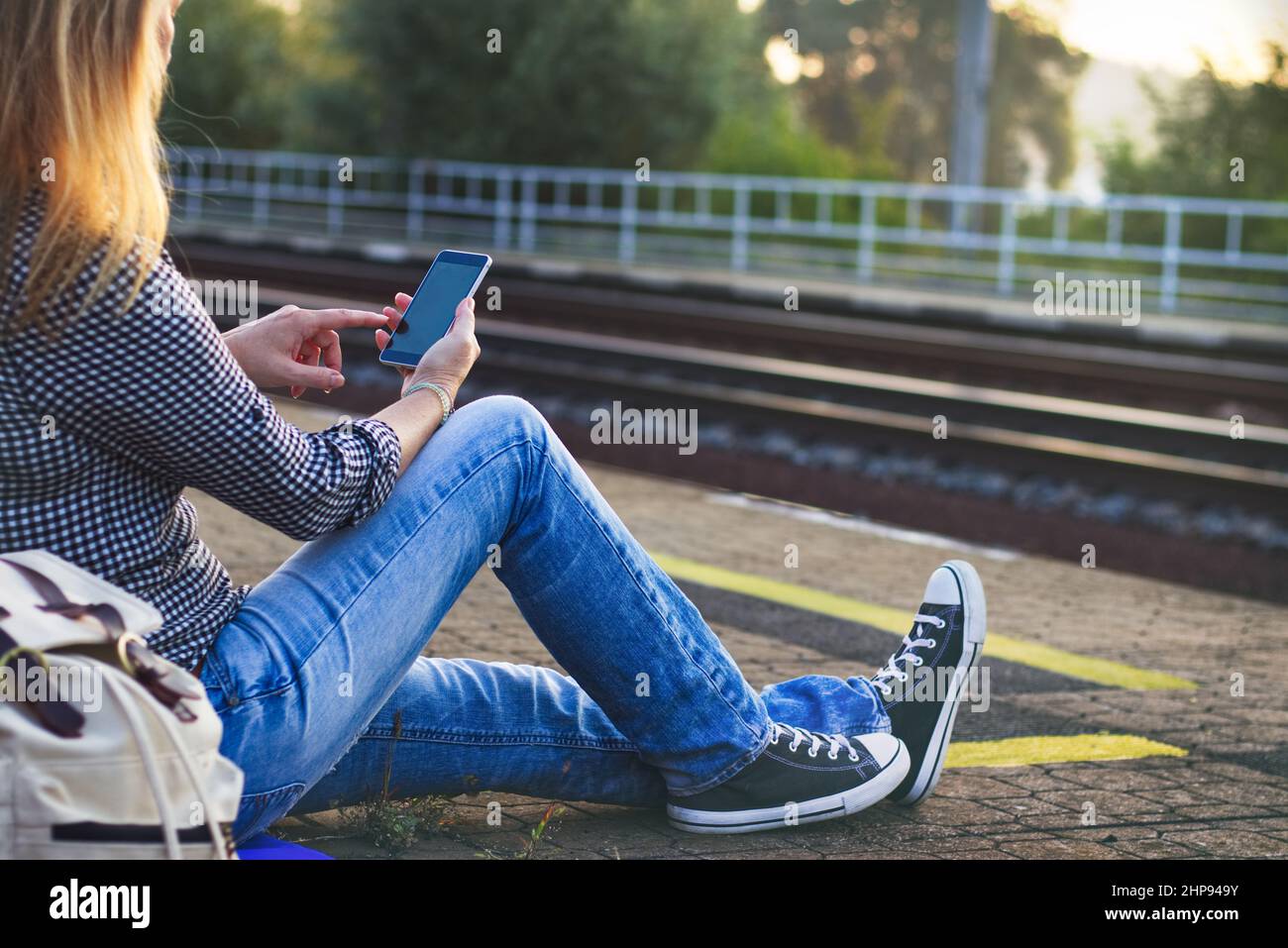 Donna che viaggia con uno smartphone durante l'attesa di un treno sulla stazione ferroviaria. Backpacker indossando jeans e sneaker Foto Stock