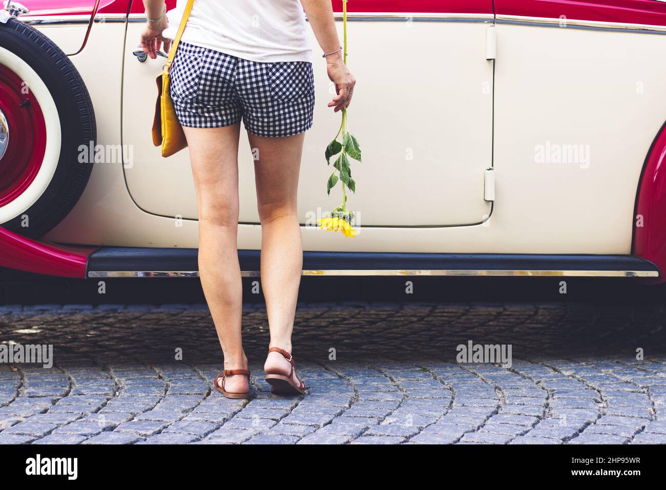 La donna indossa pantaloncini e tiene in mano il fiore, sta aprendo la porta dell'auto classica. Ragazza che indossa abiti casual viaggiando in auto retrò Foto Stock