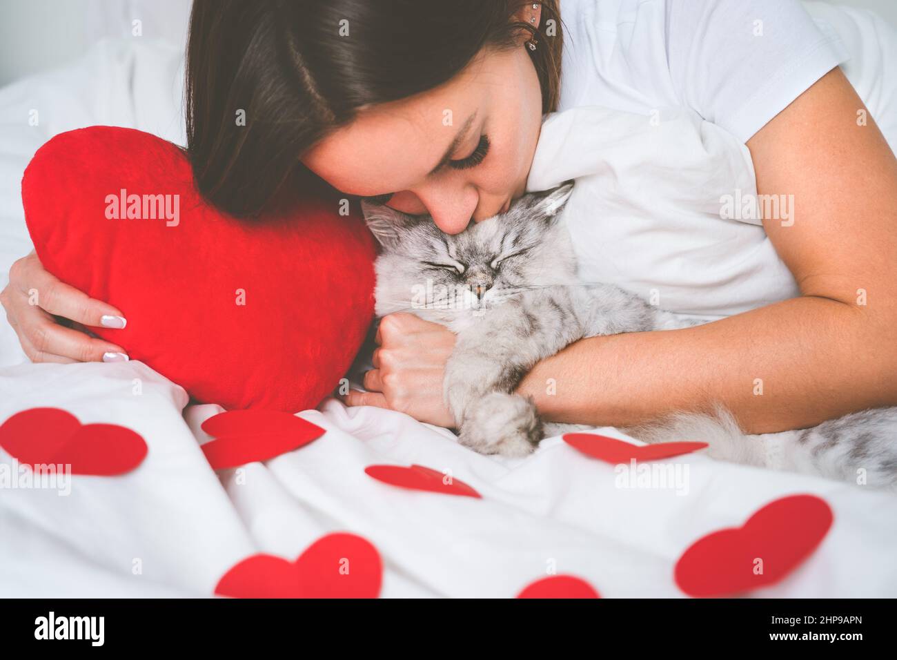 Donna sdraiata sul letto con cuori rossi e baciando il suo bel gatto. Amore per gli animali domestici. Concetto di San Valentino Foto Stock