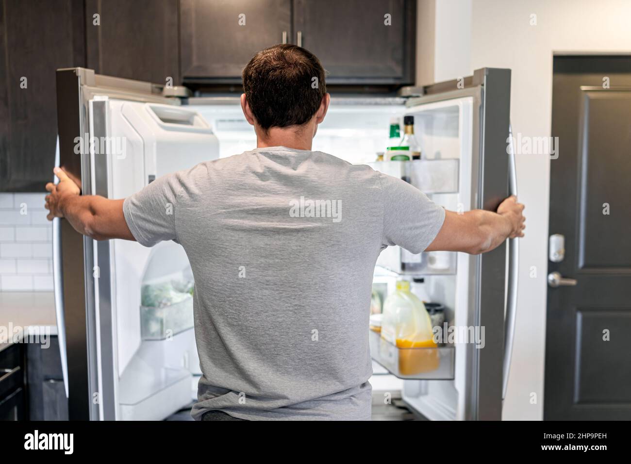Retro di uomo affamato aprire frigorifero porte elettrodomestici frigorifero alla ricerca di cibo all'interno con condimenti e succo di frutta in cucina moderna Foto Stock