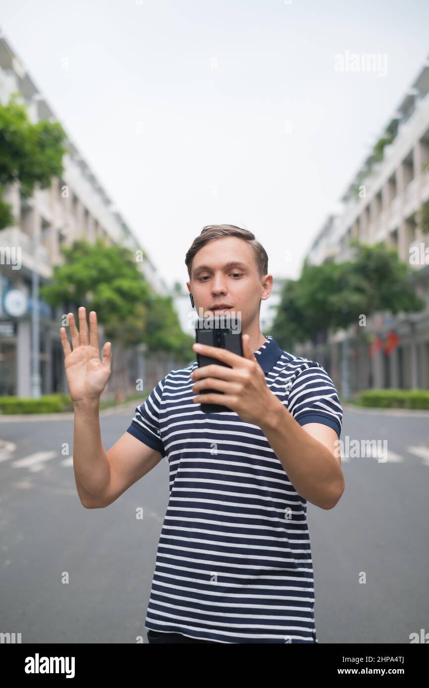 Il giovane caucasico cammina lungo la strada e effettua videochiamate al telefono con cuffie wireless. Utilizzando lo smartphone. Foto di alta qualità Foto Stock