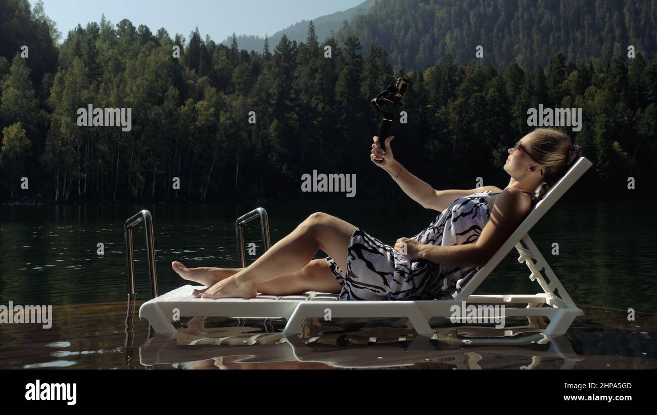 Donna che scatta con la stabilizzazione gimbal a pellicola portatile per smartphone. Ragazza riposo, relax, sdraiati sul molo in occhiali da sole, fare selfie. Signora blogger bro Foto Stock