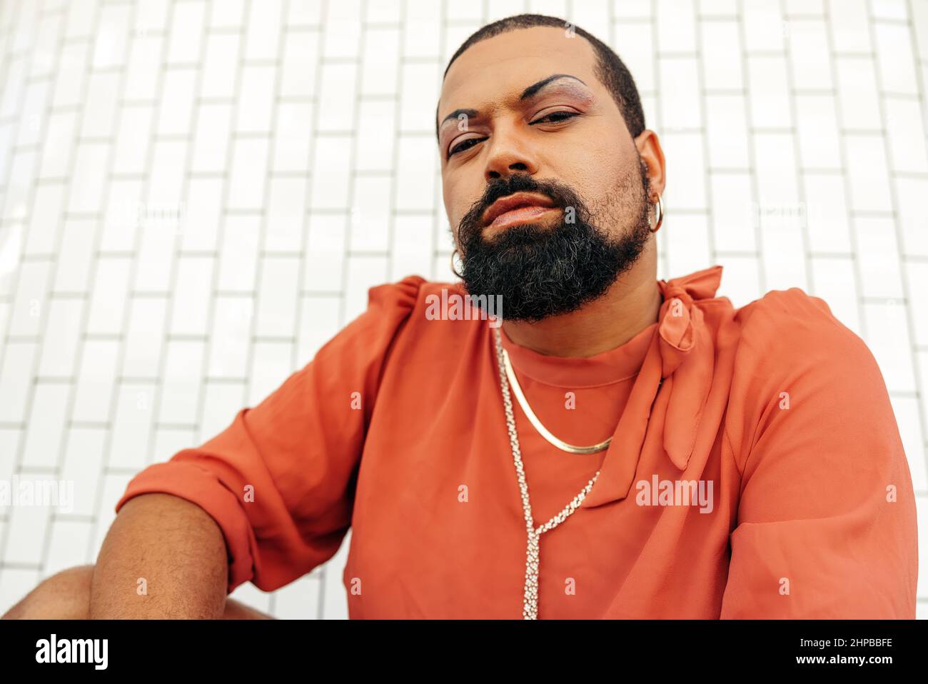 Giovane e sicura regina di trascinamento che guarda la fotocamera mentre squatting su uno sfondo bianco. Giovane uomo non conforme che indossa il make up e gioielli. SÌ Foto Stock
