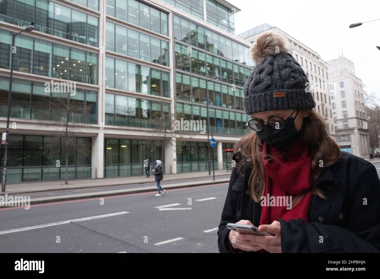 Donna che indossa cappello di lana, occhiali e maschera facciale utilizzando il telefono cellulare su una strada del Regno Unito di Londra Foto Stock