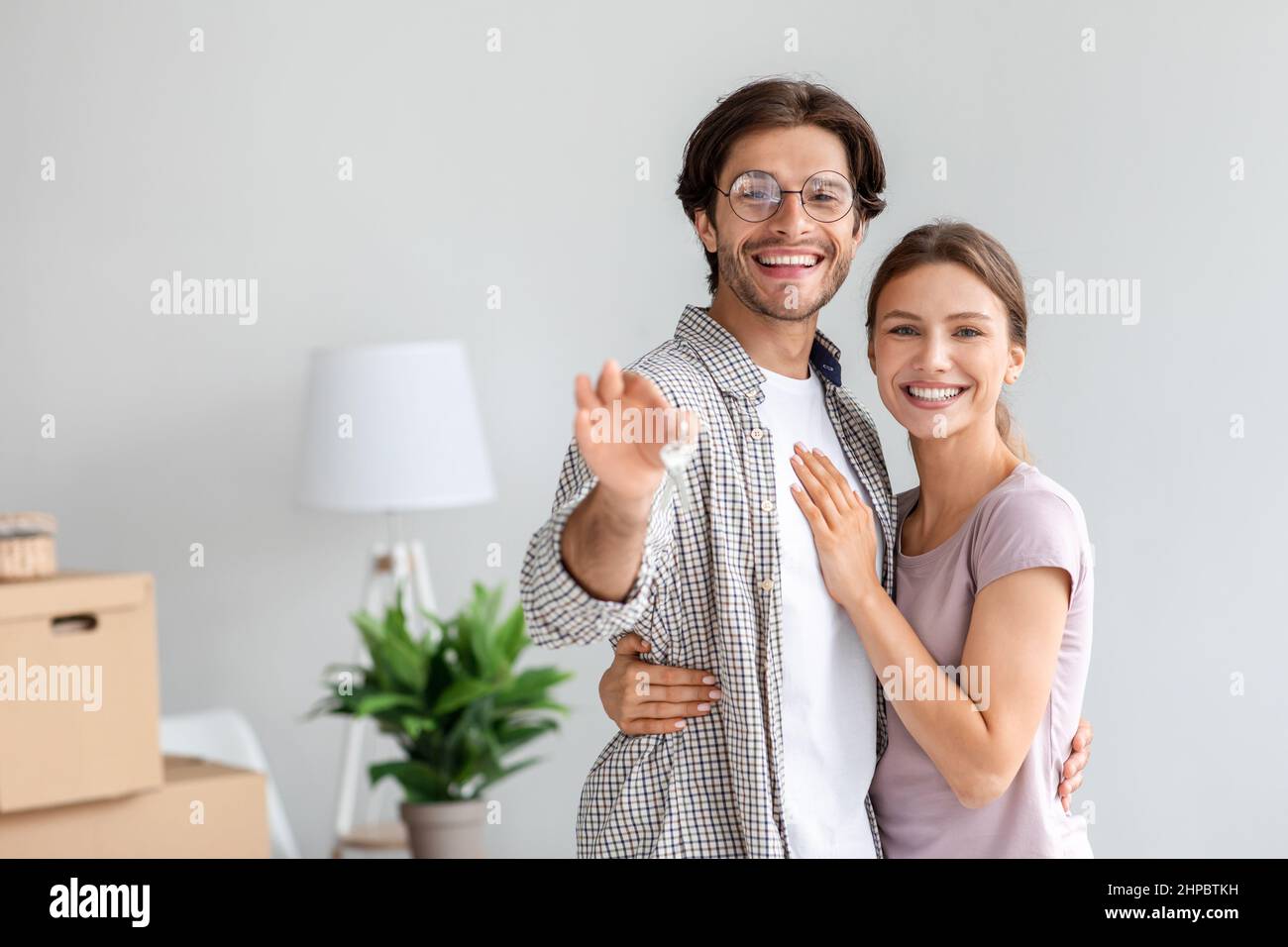 Il giovane caucasico soddisfatto moglie e marito in bicchieri mostra chiavi in proprio appartamento in camera con scatole di cartone imballato Foto Stock