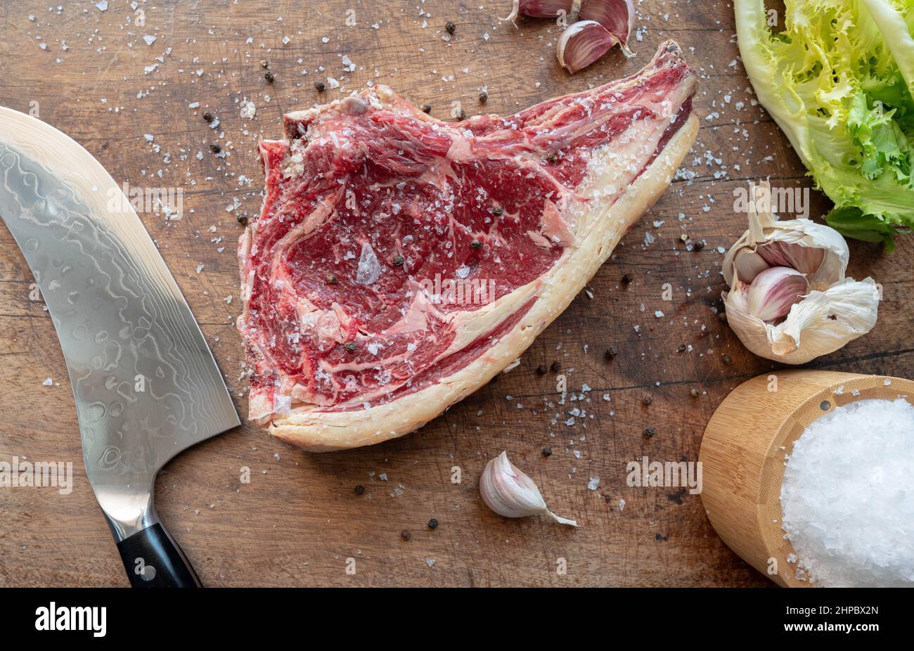 Deliziosa bistecca di manzo cruda rubia gallega stagionata su tavola di legno con coltello, insalata verde e condita con scaglie di sale marino, pepe, aglio Foto Stock