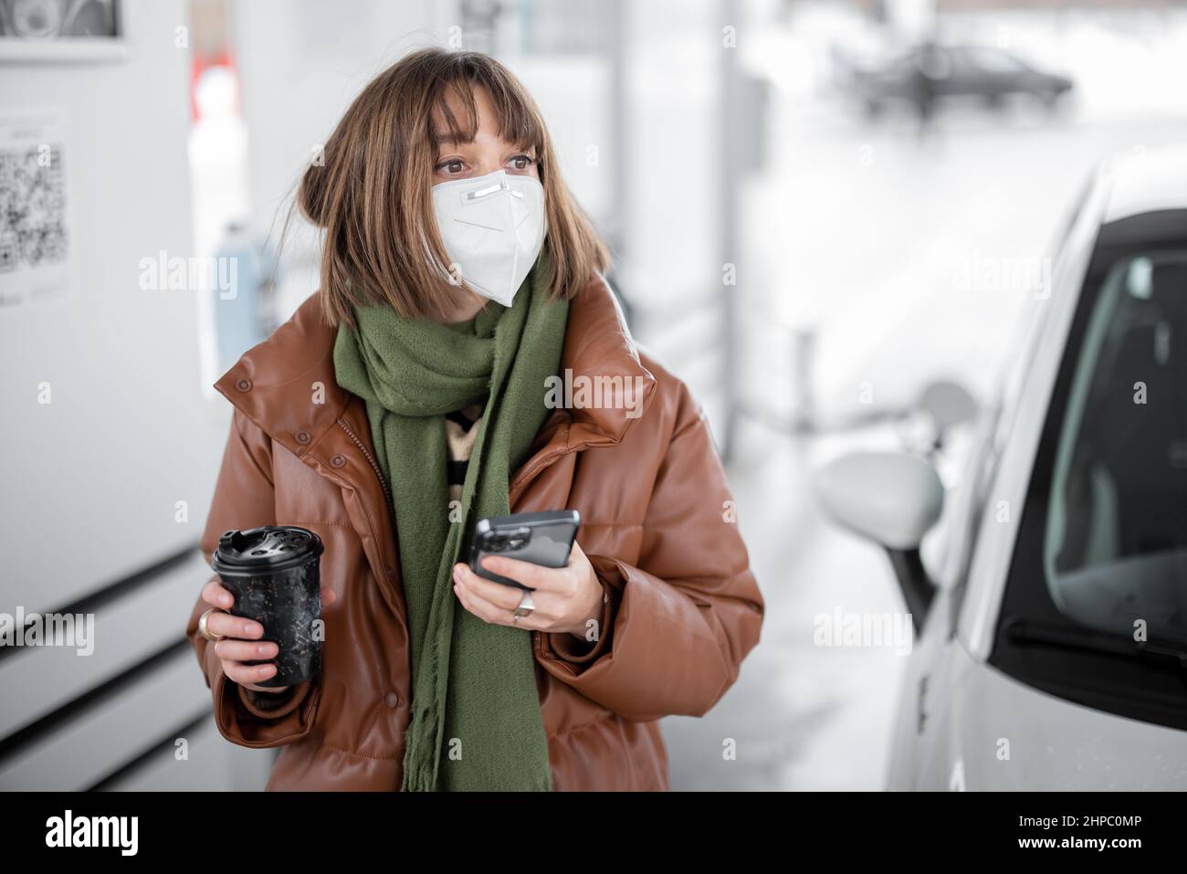 Donna in maschera di faccia che tiene smartphone e caffè tazza mentre si rifornisce l'auto al distributore di benzina. Foto Stock