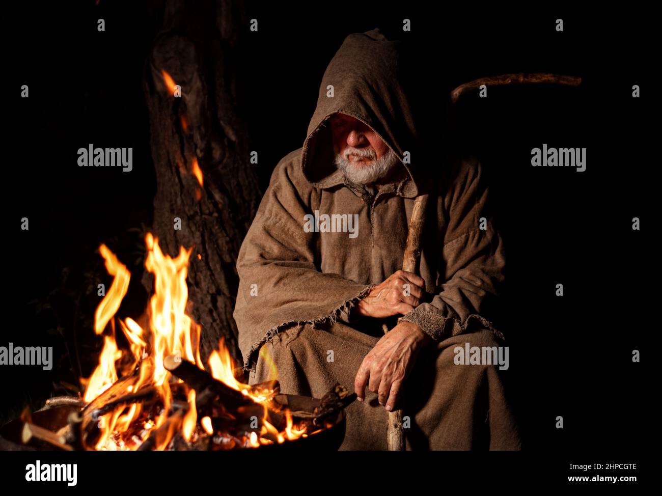 Vecchio povero uomo medievale pensieroso seduto accanto al fuoco Foto Stock