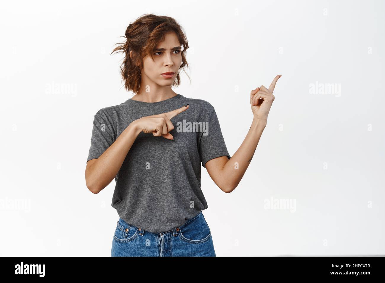 Ritratto di ragazza carina con capelli ricci, lo studente sembra confuso quando punta le dita a destra, vedere smth strano, puzzle, si erge su sfondo bianco Foto Stock