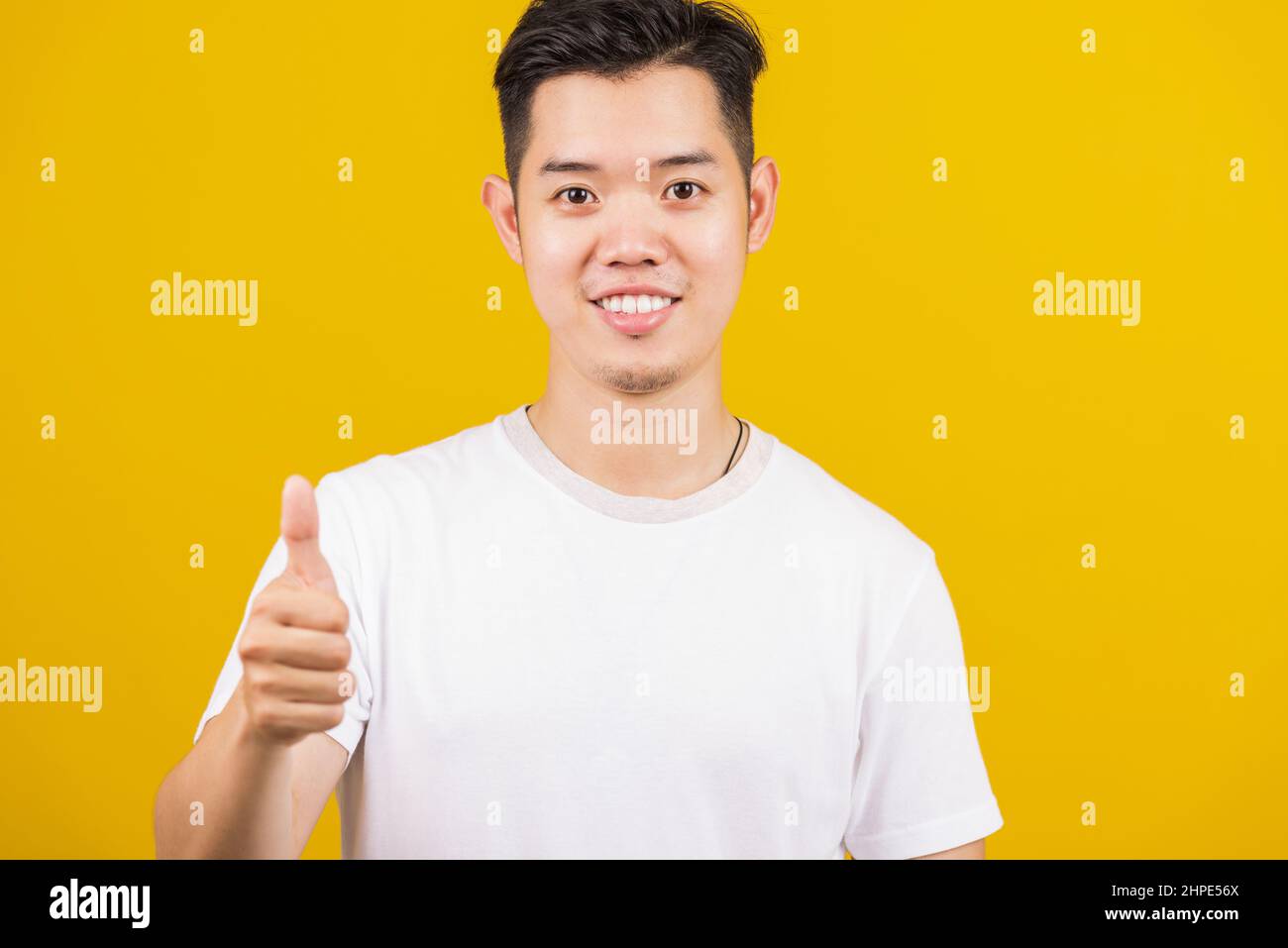 Asiatico bel giovane uomo sorridente positivo mostrando pollici verso l'alto gesto buono o come segno guardando alla macchina fotografica, studio girato isolato su sfondo giallo Foto Stock