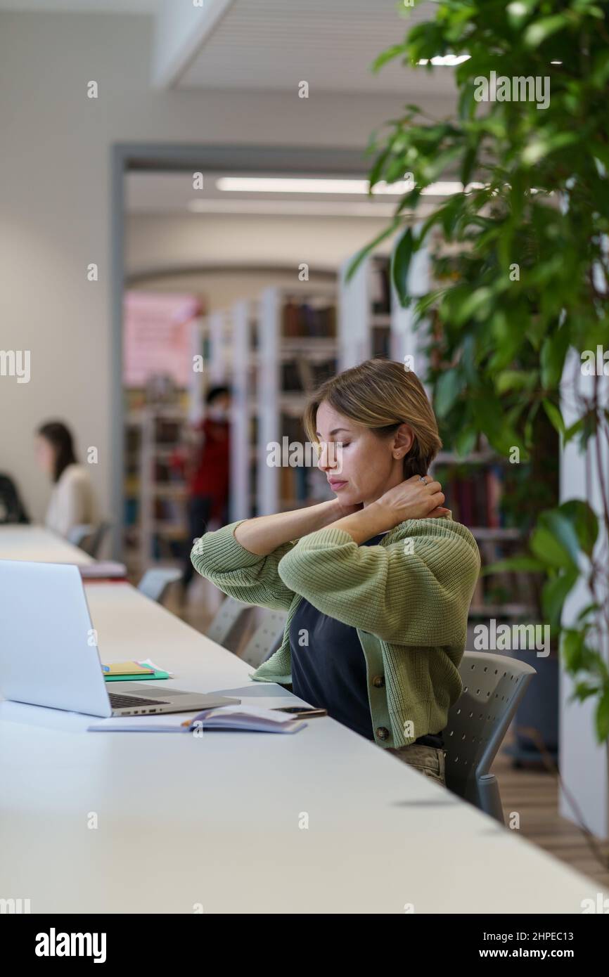 Donna stanca massaggiando il collo e guardando fuori dalla finestra mentre si lavora su un computer portatile nella biblioteca pubblica Foto Stock