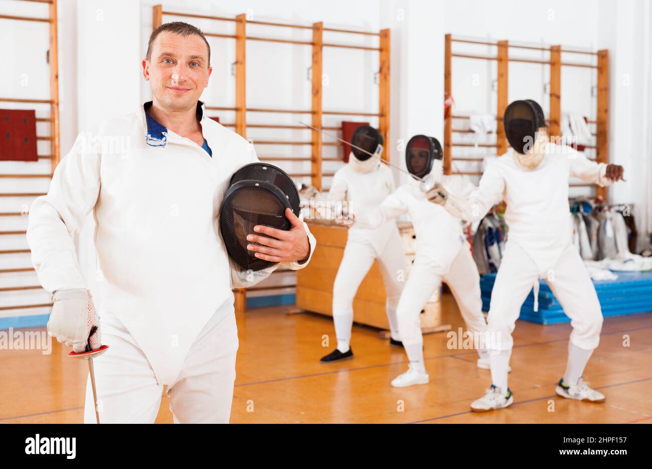 Attiva fencer maschile giovane in uniforme con maschera e lamina nella stanza della scherma Foto Stock
